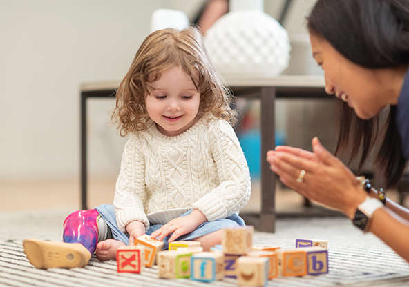 A preschool age girl with a prosthetic leg is at a medical appointment. The child is meeting with her occupational therapist. The child is sitting on the floor building with wooden toy blocks. The medical professional is sitting on the floor assisting the girl. Both individuals are smiling.